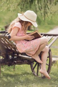 A young girl in a hat reads a book while sitting on a rustic wooden wagon outdoors.